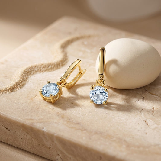Gold earrings with clear gemstones on a beige stone surface
