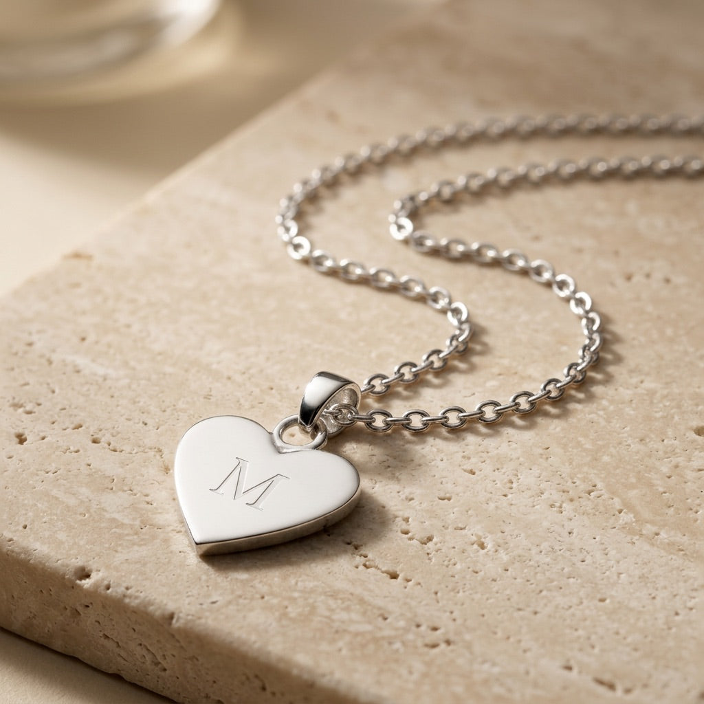 Silver heart-shaped pendant necklace with engraved letter 'M' on a beige stone surface.