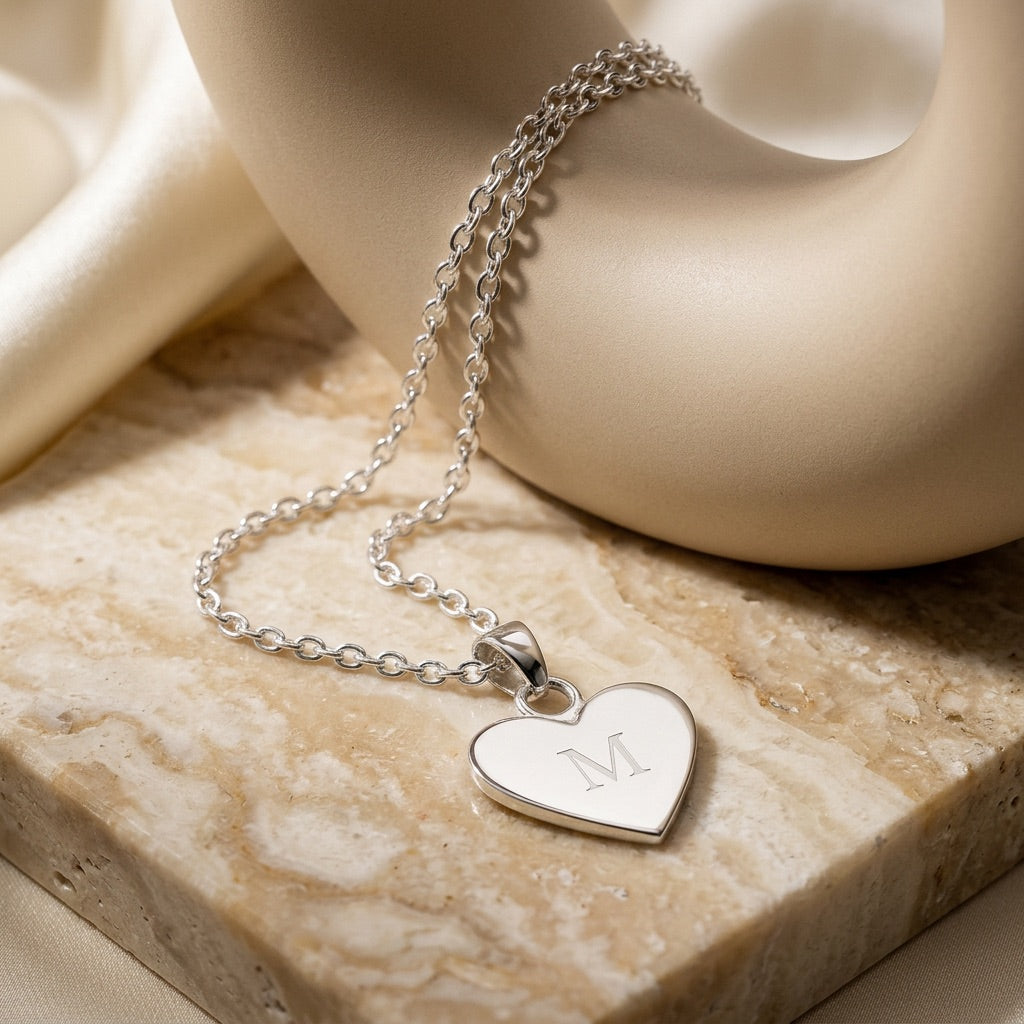 Silver heart-shaped necklace with a engraved letter 'M' on a beige stone surface.