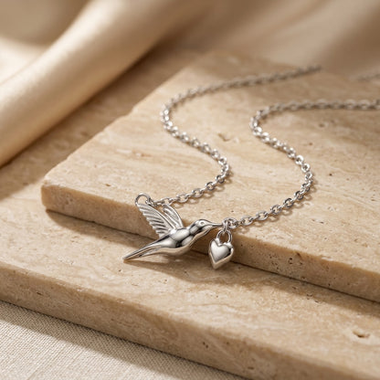 Silver necklace with a hummingbird and heart charm on a beige surface
