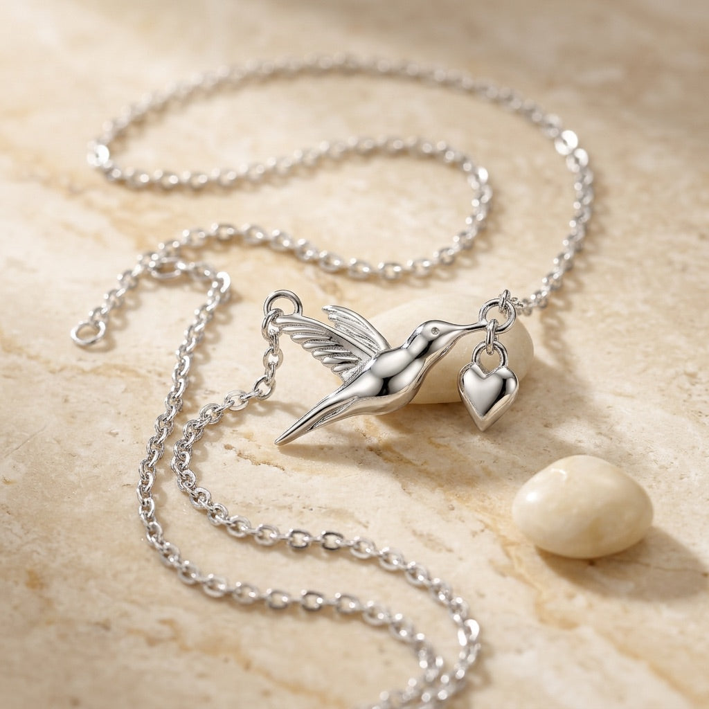 Silver necklace with a hummingbird pendant and heart charm on a beige stone surface