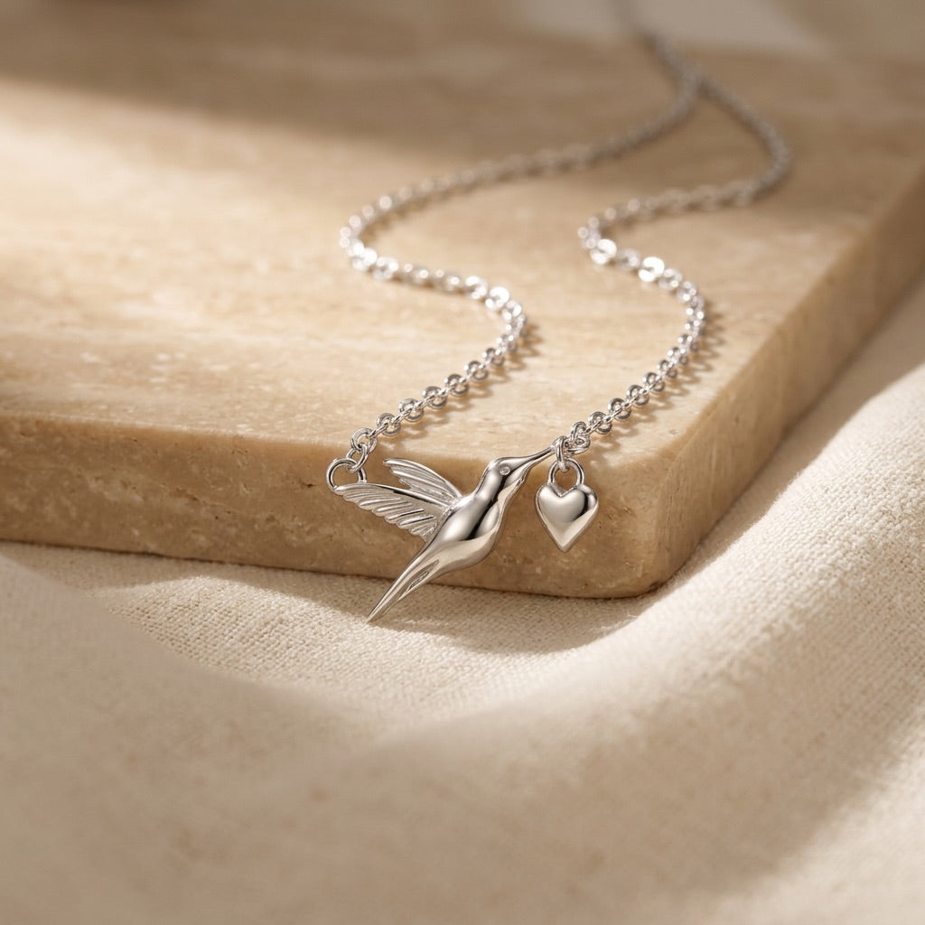 Silver necklace with a hummingbird and heart pendant on a beige surface