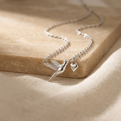 Silver necklace with a hummingbird and heart pendant on a beige surface