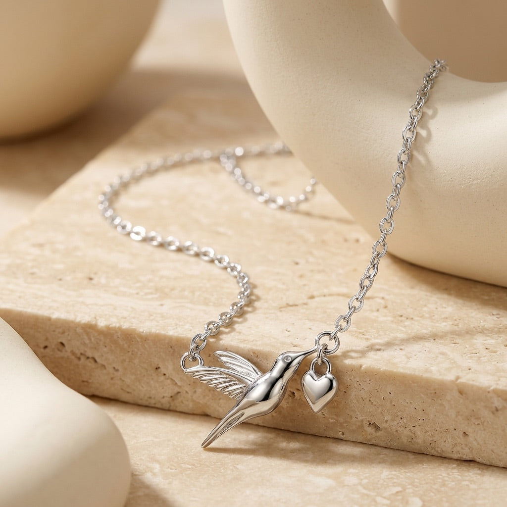 Silver necklace with a hummingbird and heart charm on a beige stone surface