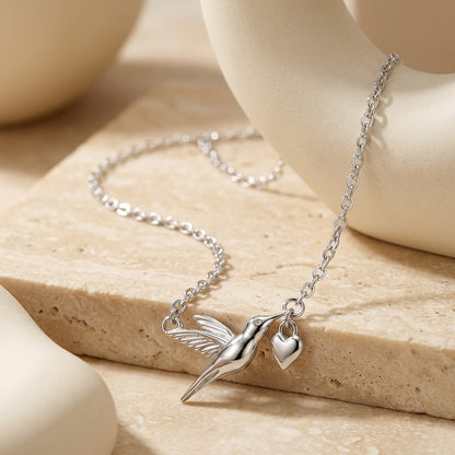 Silver necklace with a hummingbird and heart charm on a beige stone surface